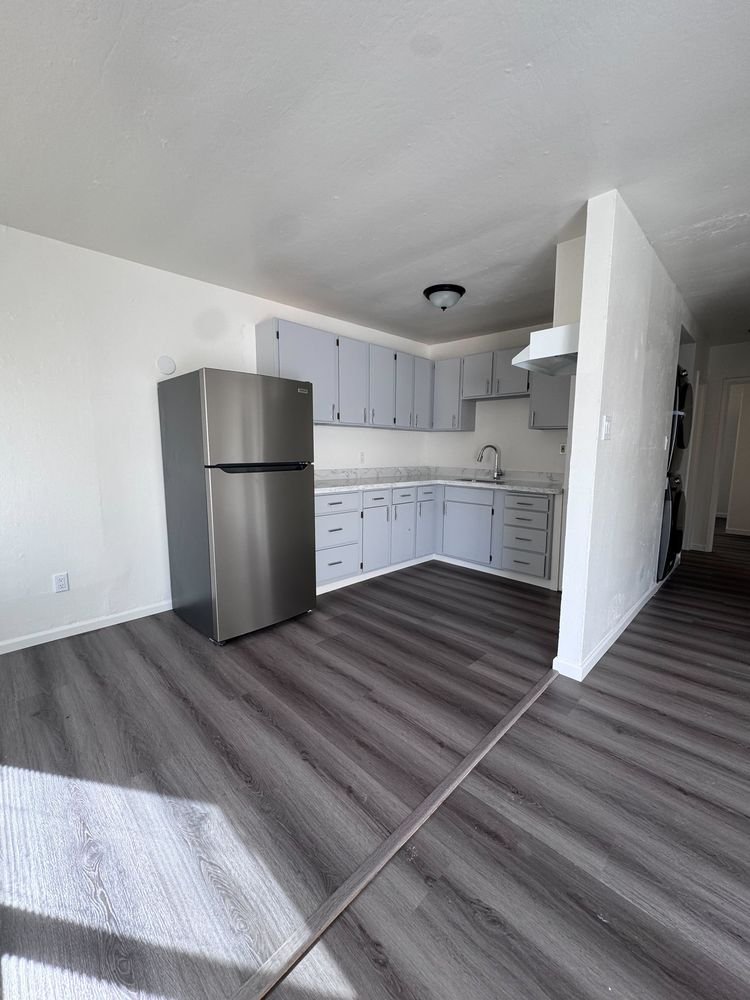 Open kitchen with gray cabinets and stainless refrigerator beside a sunny living area